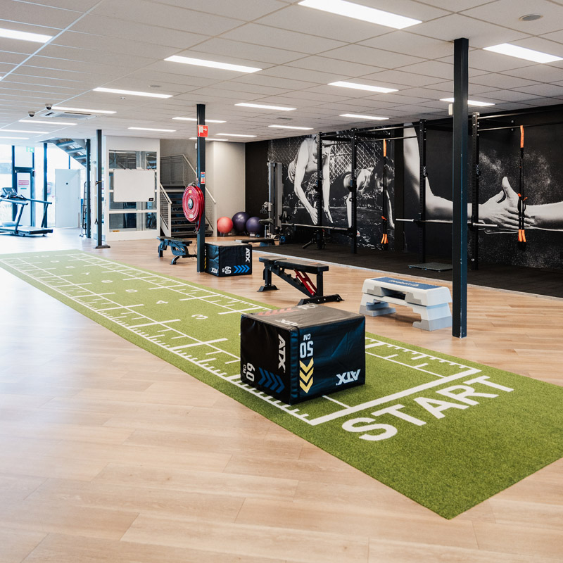 Moderne fitnessruimte of fysiopraktijk. Een lange groene kunstgrasmat met een 'START'-markering, plyoboxen en trainingsbankjes. Donkere muur met atletenafbeelding