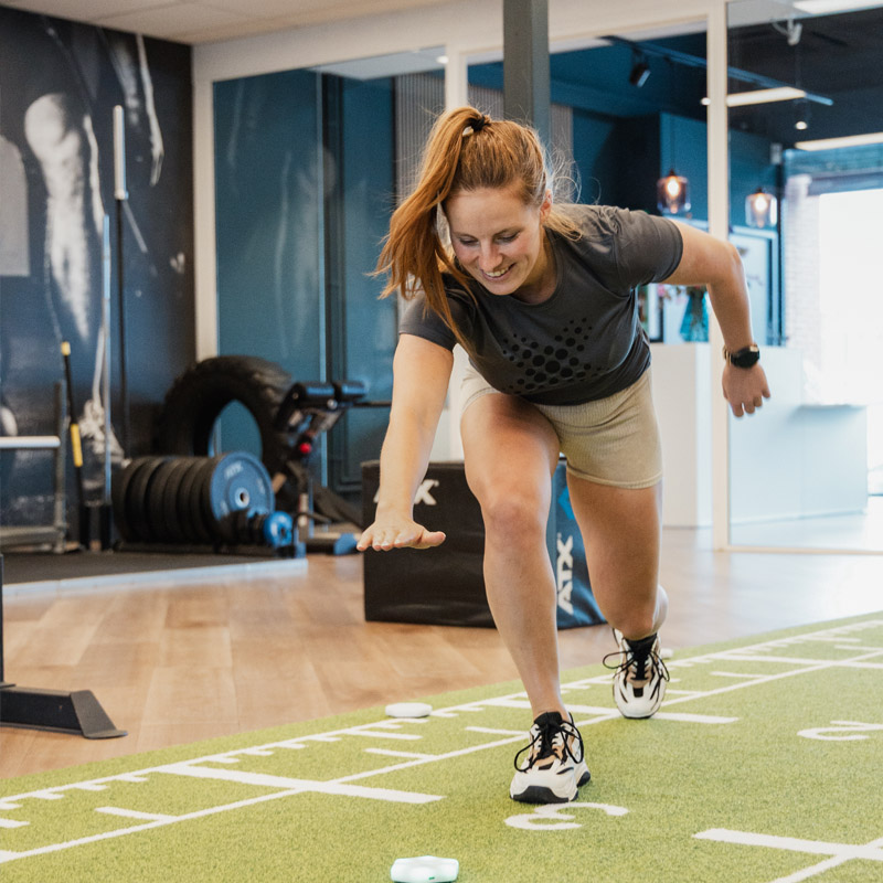 Een lachende vrouw met rood haar in een fysiotherapie/fitnessruimte doet een strekoefening op een groene sportmat. Ze reikt naar voren terwijl ze een lunge doet