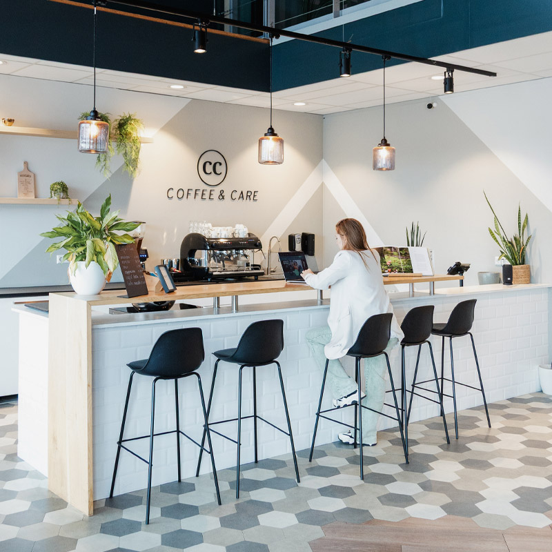 Een moderne koffiehoek in een praktijk, met een witte bar, barkrukken en een espresso-apparaat. Een vrouw werkt aan een laptop terwijl ze op een kruk zit