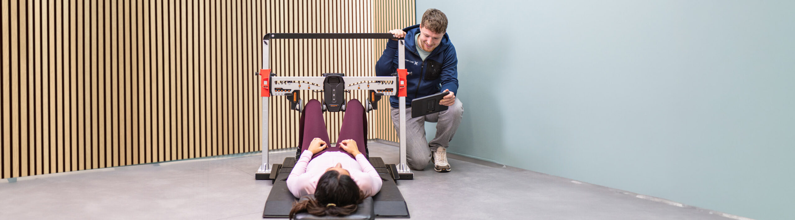Een vrouw ligt op een trainingsapparaat terwijl een fysiotherapeut haar voortgang volgt op een tablet. Ze traint haar benen met het apparaat, dat boven haar knieën is geplaatst.