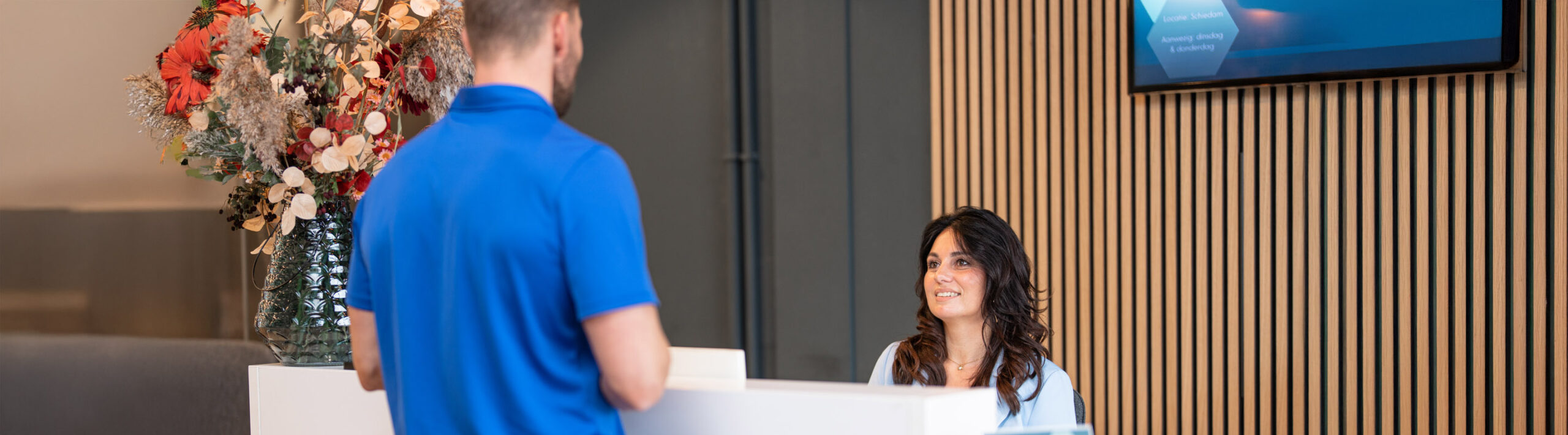 Een glimlachende receptioniste verwelkomt een man aan een witte balie in de Joint.Care fysiotherapie praktijk in Maassluis.